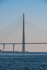 Obraz premium Modern cable-stayed bridge spans across a calm body of water with clear blue sky and distant land visible in the background