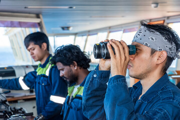 Obraz premium Professional ship crew focused on bridge control systems as one deck officer conducts visual lookout with binoculars to ensure safe navigation at sea.