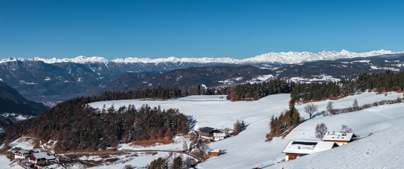 Snow covered meadows with chalet style buildings and barns sit below jagged Dolomites peaks in northern Italy, daylight midwinter, crisp light and long shadows. © Aerial Film Studio