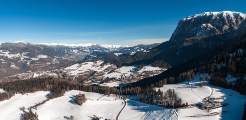 Aerial view of snow dusted Dolomites in northern Italy, a sheer faced massif at right, alpine meadows and hamlets below, a chalet hotel on a sunlit slope at midday.