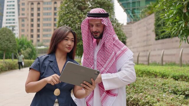 Asian businesswoman and Arab Miiddle eastern businessman talking about cross-border and international business, walking along downtown park.