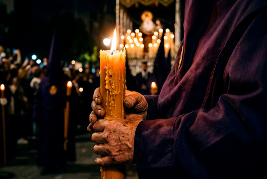 Close-up of hands holding a large burning candle during a Semana Santa procession in Spain. A traditional robe sleeve is visible while warm candlelight illuminates melting wax
