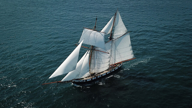 Plougonvelin, France - 12 August 2021: Aerial view of a classic sailing ship, its white sails billowing against the deep blue ocean, a dance of light and shadow on the water's surface.