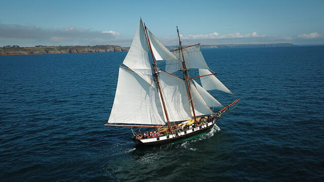 Aerial view of the majestic sailing ship La Recouvrance with billowing white sails glides across the deep blue waters near the rocky cliffs of Pointe Saint Mathieu, Plougonvelin, Brittany, France.