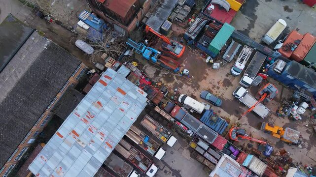 Drone view of vehicle scrap metal yard, diagonal top-down reverse flight over heavy machinery, stacked containers, parked cars and industrial clutter at a recycling facility.