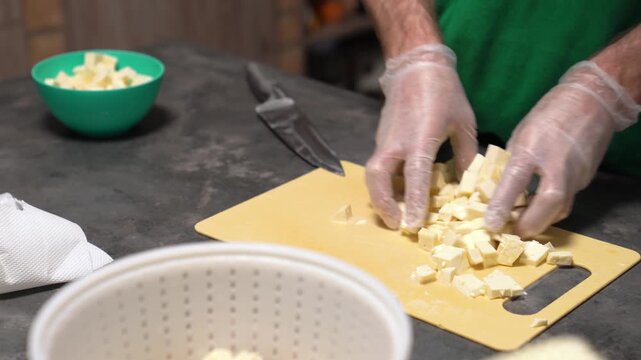 Person wearing gloves preparing food, cutting a block of white cheese or tofu into cubes