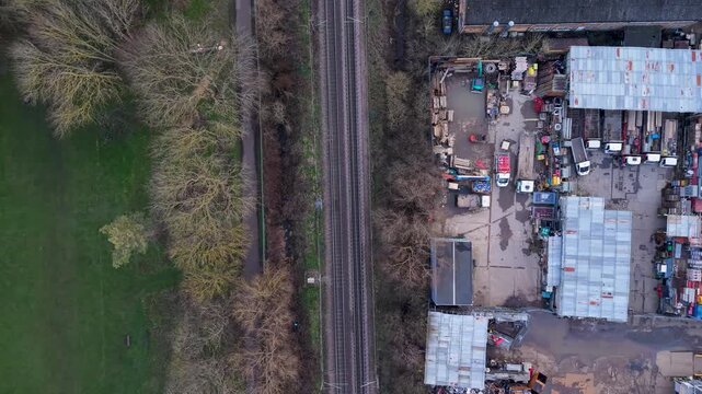 Drone pan over grass and trees, crossing railway tracks into a car breakers yard with rusted roofs and abandoned vehicles, showing stark rural-to-urban transition.