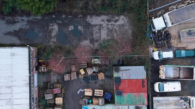 Fox approaches industrial yard as top-down drone reverses over shipping containers, pallets and parked trucks, showing urban wildlife entering a gritty logistics area.