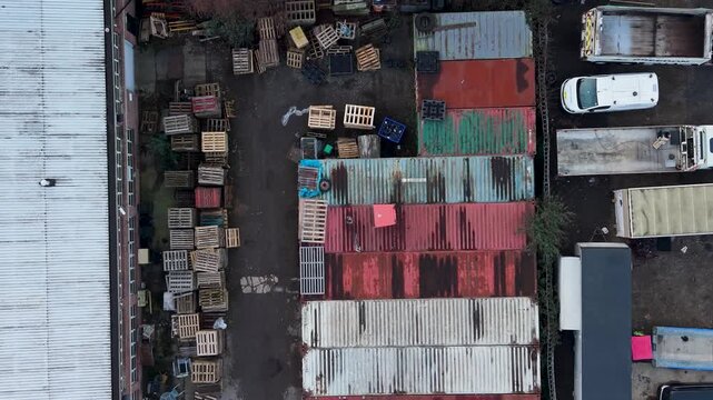 Drone top-down shipping containers in industrial yard, reversing over colorful rows beside warehouse roof, parked trucks, vans, and old vehicles in a busy logistics depot.