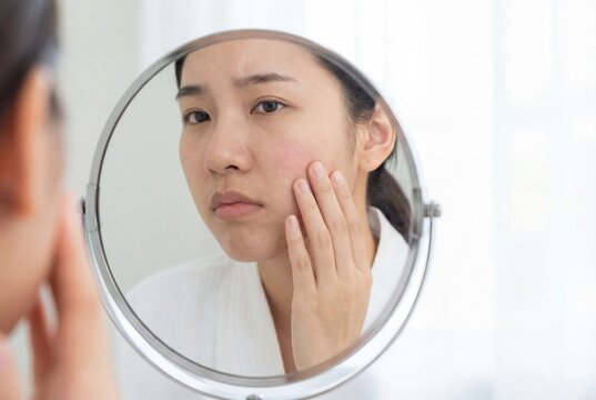 Asian woman looking at redness on her cheek in a mirror reflection