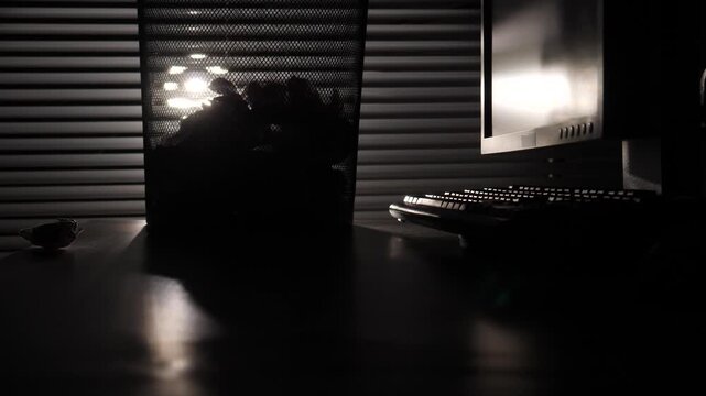 Office desk showcasing a silhouette of a wastebasket overflowing with crumpled paper, next to a computer monitor and keyboard, illuminated by light filtering through blinds in a dark room