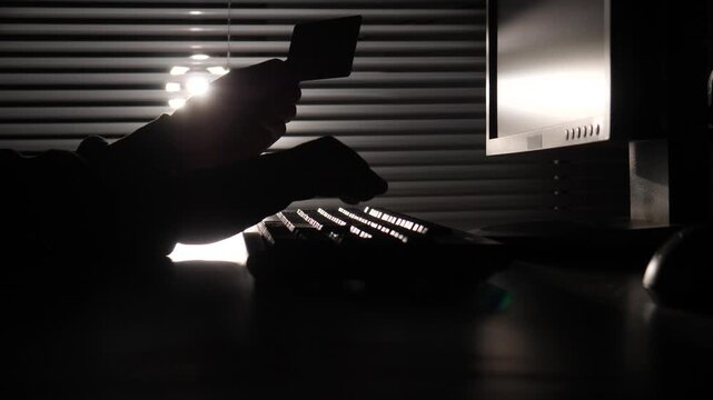 Silhouette of a fraudster's hands typing on a computer keyboard while holding a credit card, emphasizing concepts of cybercrime, data theft, and online banking security