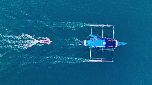 Blue outrigger boat towing small pink vessel across deep teal water