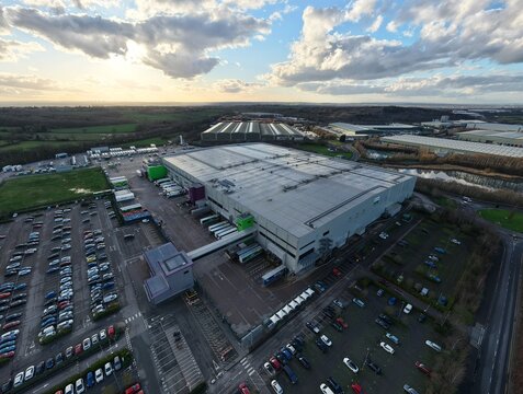 Tamworth, United Kingdom - 21 February 2026: Aerial view of a vast distribution center casting long shadows, with orderly rows of parked cars contrasting with the distant, wilder green landscape.