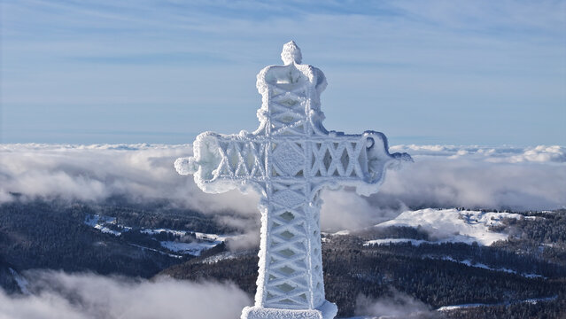Aerial view of the ice-encrusted cross atop Le Reculet mountain, a beacon against the backdrop of misty valleys and a winter sky, Thoiry, Auvergne-Rhone-Alpes, France.