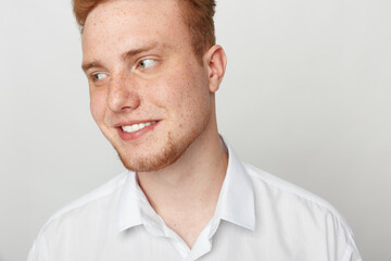 Obraz premium Young redhead man with freckles smiles looking sideways wearing white shirt on light background. Concept of casual style, happiness, and natural male portrait in studio lighting.