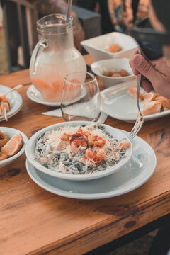 Primer plano de persona comiendo pasta con camarones en una mesa de restaurante al aire libre con bebidas y entradas