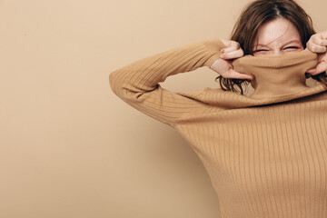 Young woman in brown sweater pulls collar over face with eyes closed, expressing playful mood on...