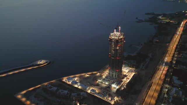 Aerial drone footage focused on a skyscraper under construction during blue hour. High-rise tower development with cranes and structural framework rising above a large urban redevelopment area
