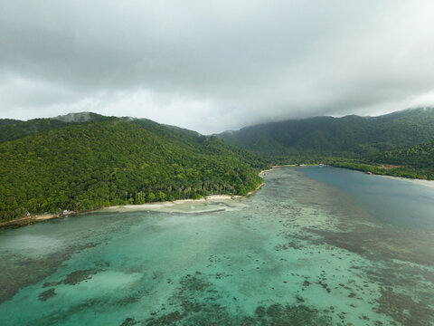 Aerial view of turquoise waters meet lush green hills under a blanket of soft clouds at Bobby Beach, Karimunjawa, Central Java, Indonesia.
