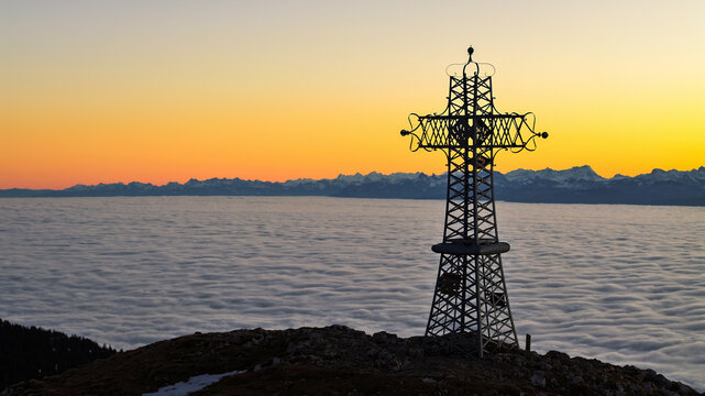Aerial view of the cross of Le Reculet standing boldly against a sea of clouds, kissed by the warm hues of the setting sun, Thoiry, Auvergne-Rhone-Alpes, France.