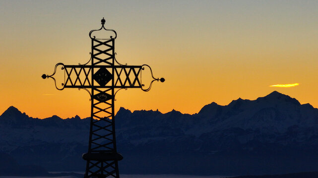 Aerial view of the cross atop Le Reculet mountain with the Jura mountain range silhouetted against the vibrant orange glow of the setting sun., Thoiry, Auvergne-Rhone-Alpes, France.
