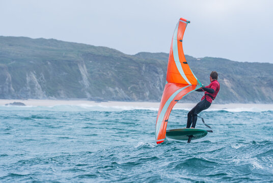 A man wing foils at sea using a bright orange hand held inflatable wing, riding a hydrofoil surfboard. Green sea, cloudy blue sky.	 Coastal cliffs in the background.