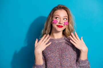 Playful young woman with pink stickers on cheeks wearing a purple sweater against a bright blue...