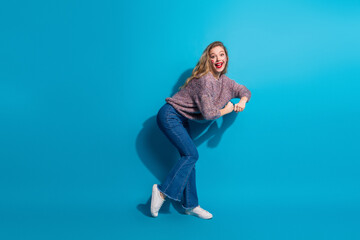 Young stylish woman posed playfully against a blue background wearing a cozy sweater and jeans for...