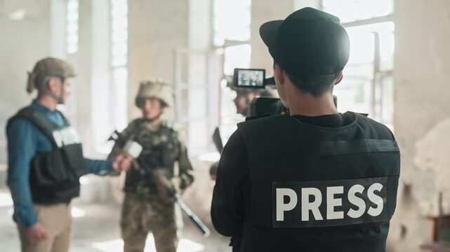Rear view of cameraman wearing bulletproof and safety helmet filming report in smoky building. Correspondent communicating on camera with military people indoor. TV reportage. Filming news.