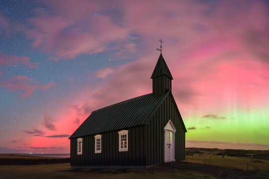 View of a black church standing against a vibrant aurora borealis sky, its simple architecture stark against the celestial dance of colors, Snaefellsnes Peninsula, Eyja- og Miklaholtshreppur, Iceland.