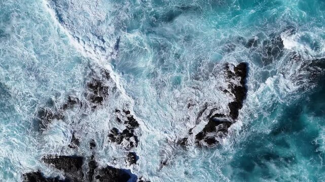 Turquoise ocean waves crash against rocky shoreline, creating foam and splashes. Aerial view to dynamic movement of water's surface. Beautiful colorful sea landscape. Close up