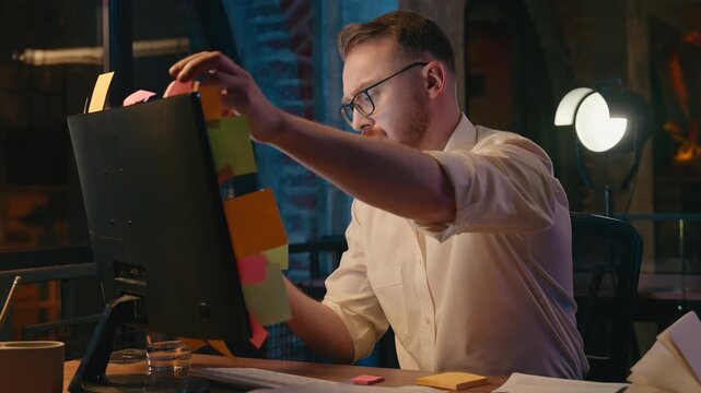 Tired Caucasian man business in night office managing sticky notes on monitor computer screen throwing reminders away confused businessman male guy in dark evening office check information on stickers