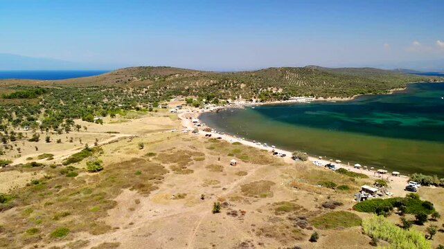 Aerial view of the beach with people, cars, and an island with green trees, contrasting with the blue sea, Cunda Island, Ayvalik, Balikesir, Turkey.