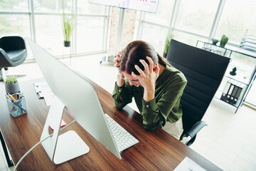 Stressed businesswoman at desk holding her head in a modern office with computer and paperwork,...