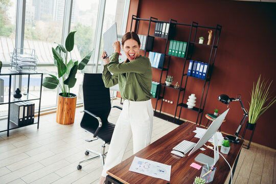 Frustrated office worker raising clipboard in anger at desk in modern workplace, stressed professional reacts to work pressure in open-plan bright office