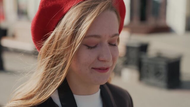 Young white woman red beret smiling on phone at bench, relaxed urban pause with soft sunlight, cozy coat and easy conversation, gentle breeze through hair and thoughtful expression