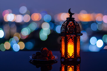 Selective focus of lantern that have moon symbol on top and small plate of dates fruit with city bokeh light background for the Muslim feast of the holy month of Ramadan Kareem.
