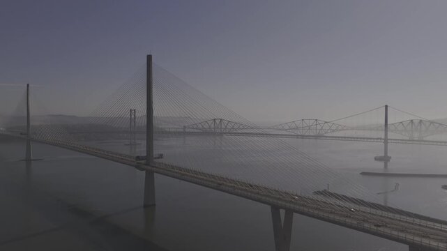 Aerial view of the three Forth Bridges spanning the Firth of Forth under a muted sky, creating a striking contrast with the calm waters, South Queensferry, United Kingdom.