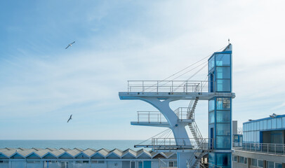 Minimalist high-dive platform stands against a clear blue sky over the sea. Seagulls soar near the architectural structure of an outdoor coastal swimming complex © John_Doo78
