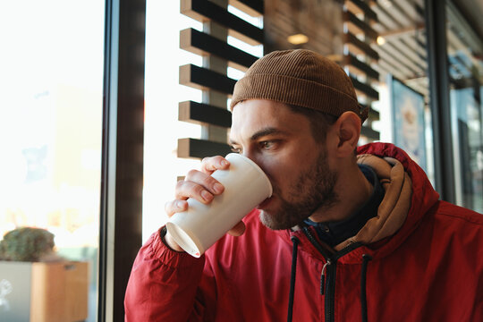 Young man in red jacket drinking carbonated soda from paper cup while sitting near window in fast food restaurant. Concept of quick meal, urban lifestyle and casual dining