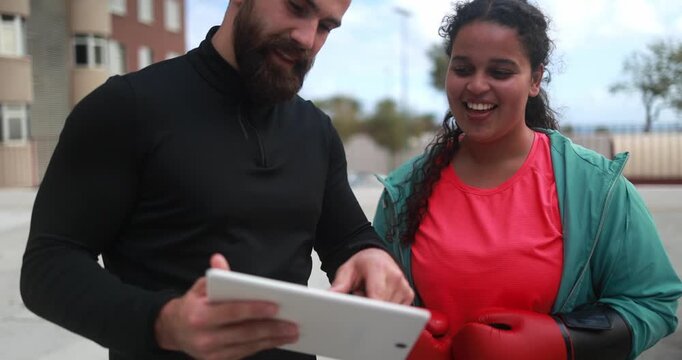 Happy curvy african american girl talking with her personal trainer while checking workout plan on tablet outdoor - Sport, healthy lifestyle and plus size people concept