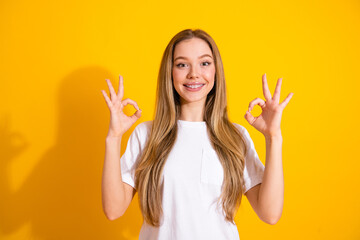 Happy young woman with braces smiles against a bright yellow background and shows an ok gesture
