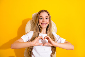 Young woman smiling with braces making a heart gesture against bright yellow background