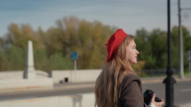 White woman photographer in red beret capturing urban skyline with mirrorless camera candid smiles and profile shots, long blonde hair, bridge and tree backdrop, sunny blue sky, stylish coat, travel