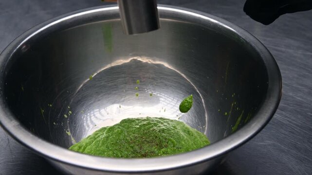Hand holding a metal grinder above a stainless steel bowl filled with vibrant green spinach mixture, showcasing the grinding process in a kitchen setting