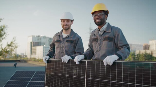 Two engineers standing on rooftop holding solar panel smiling to camera in work uniforms. Urban cityscape. Promoting renewable energy and teamwork in solar installation. Environmental and sustainable