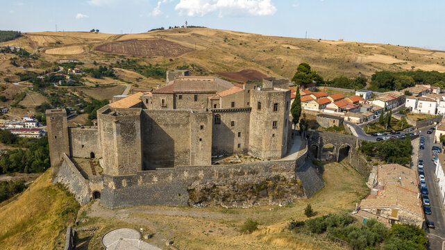 Panorama of a massive medieval stone fort with multiple towers, situated on a grassy hill overlooking a vast landscape of rolling golden plains. It's the Norman Castle of Melfi in Basilicata, Italy.