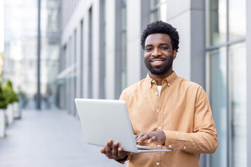 Confident young adult man standing smiling with a portable computer in front of a modern corporate...