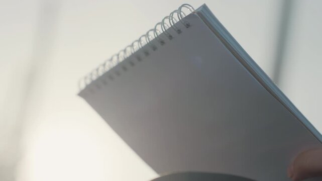 Closeup white hand holding notebook, spiral binding and empty pages backlit by soft sunlight, shallow depth of field, poised composition, quiet anticipation and minimalist mood, waiting for sketch